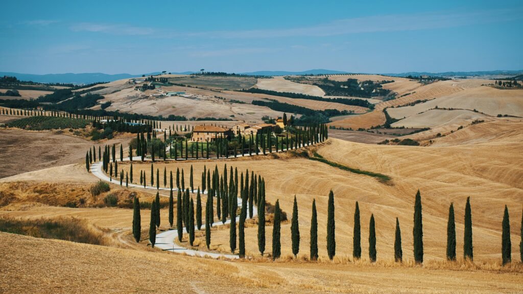 Beautiful Tuscan countryside with a winding, cypress-lined road and rustic villa under a blue sky.