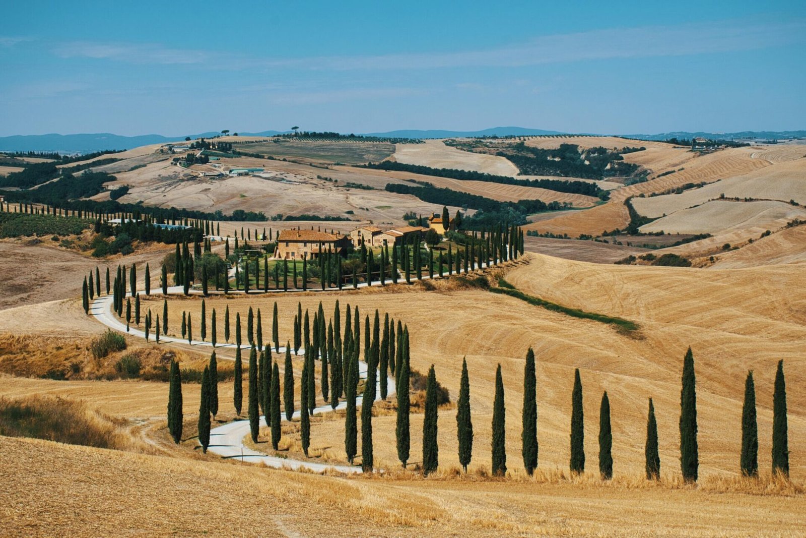 Beautiful Tuscan countryside with a winding, cypress-lined road and rustic villa under a blue sky.