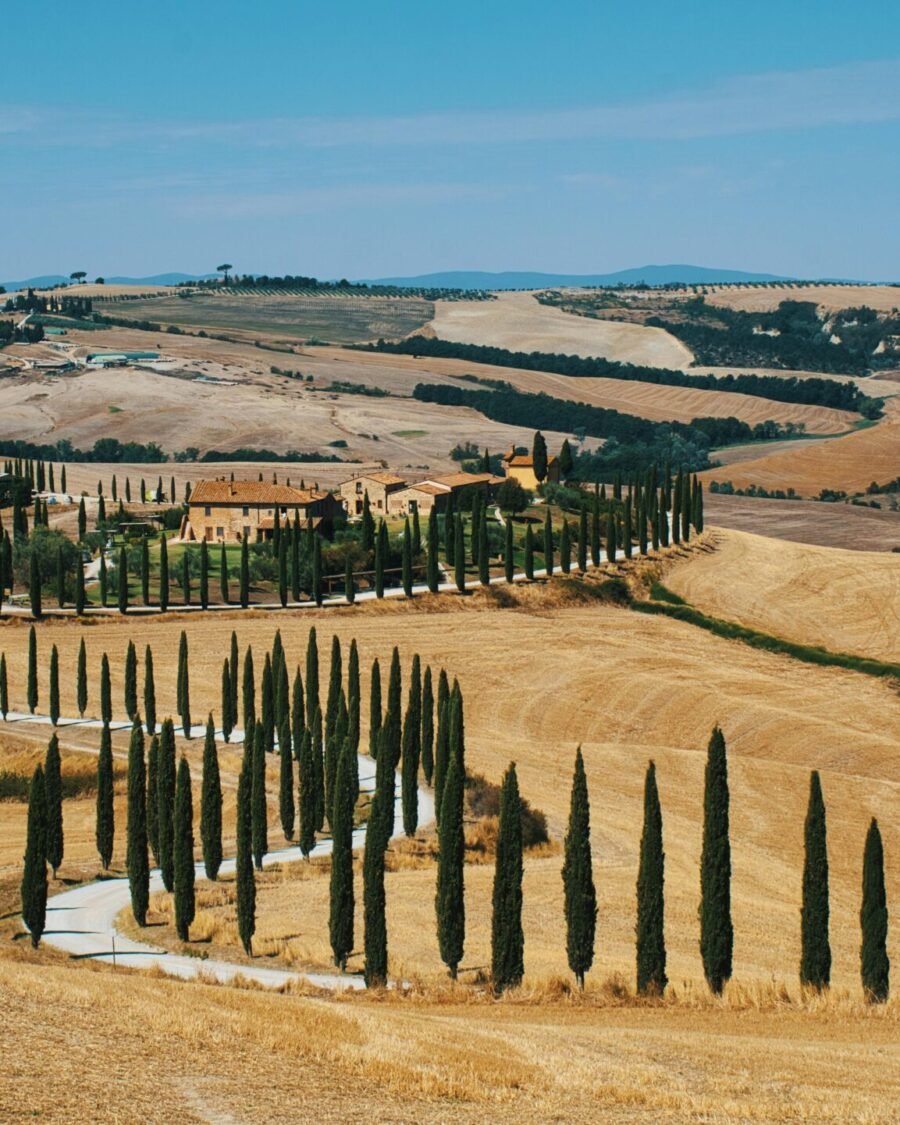 Beautiful Tuscan countryside with a winding, cypress-lined road and rustic villa under a blue sky.