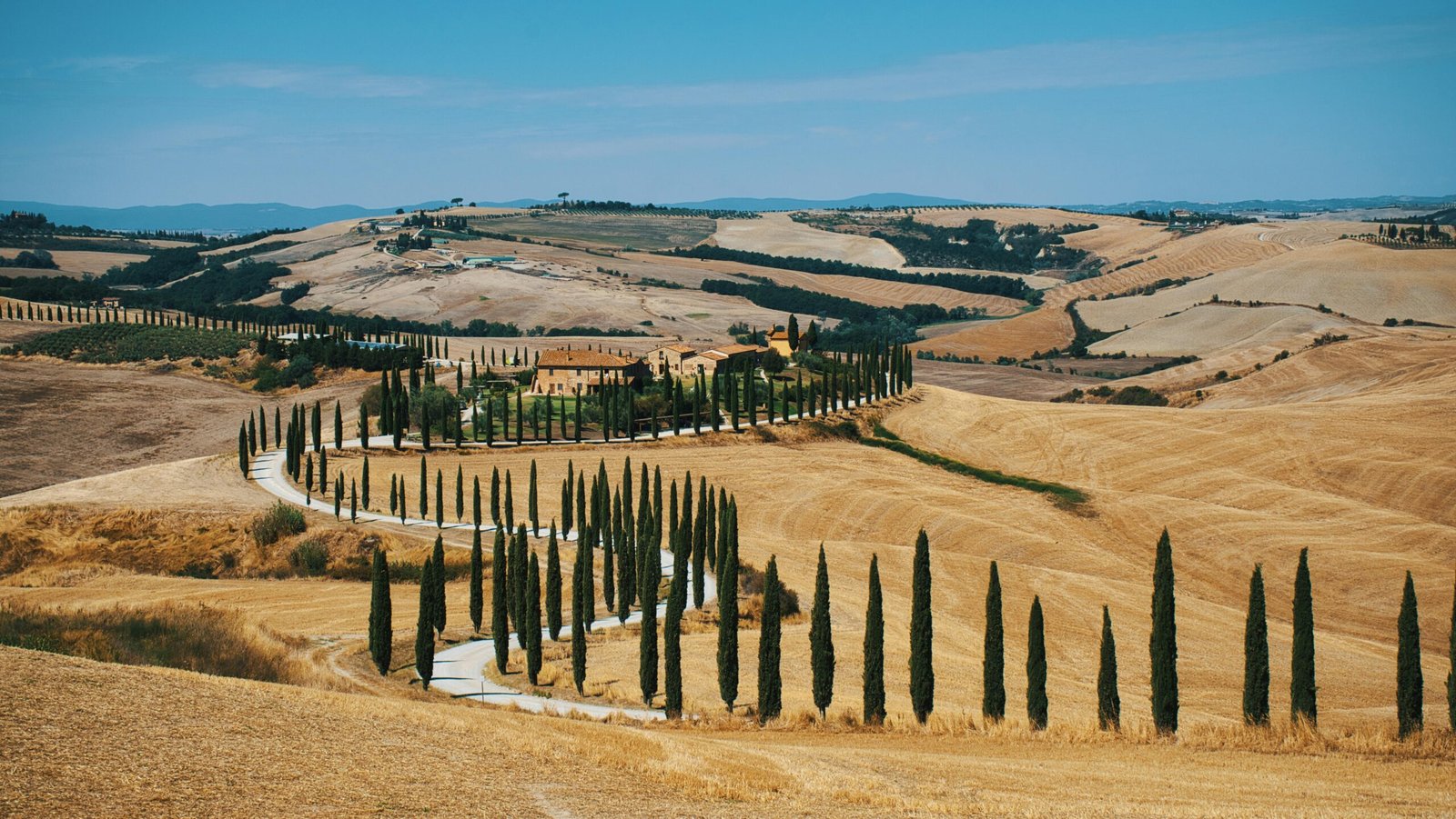 Beautiful Tuscan countryside with a winding, cypress-lined road and rustic villa under a blue sky.